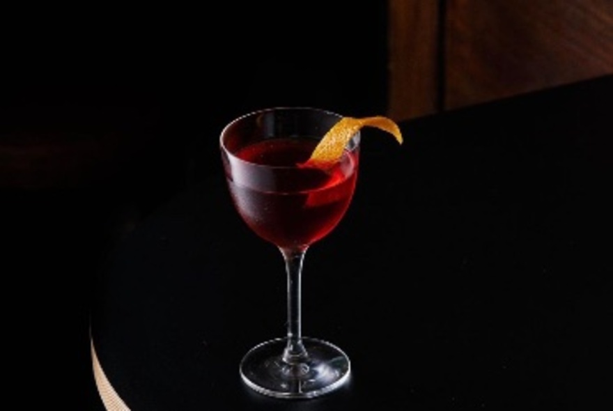 A red cocktail in a small glass with orange peel garnish. 