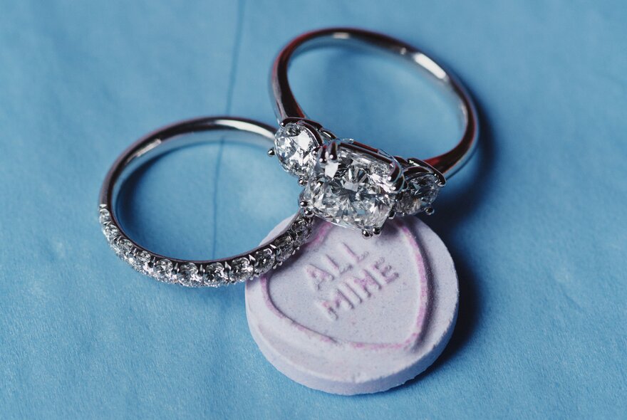 A wedding band and engagement ring set resting on a sugar lolly with a heart that reads 'All Mine'.