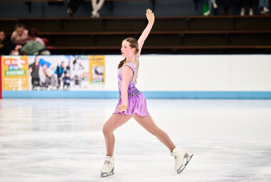 A female figure skater performing on ice in a pinkish dress, one arm stretched above her head. 