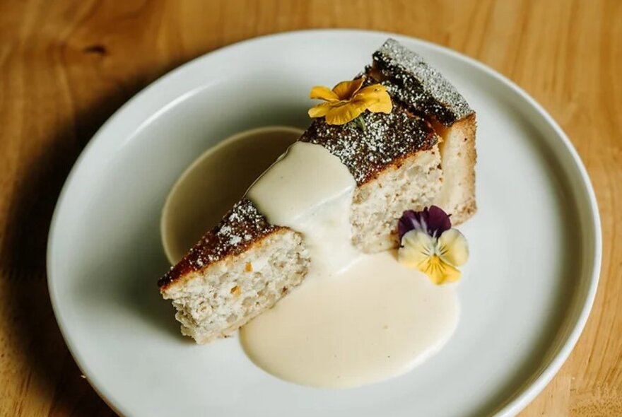 A decadent slice of cake with a white cream sauce and edible flowers as garnish, presented on a white plate.