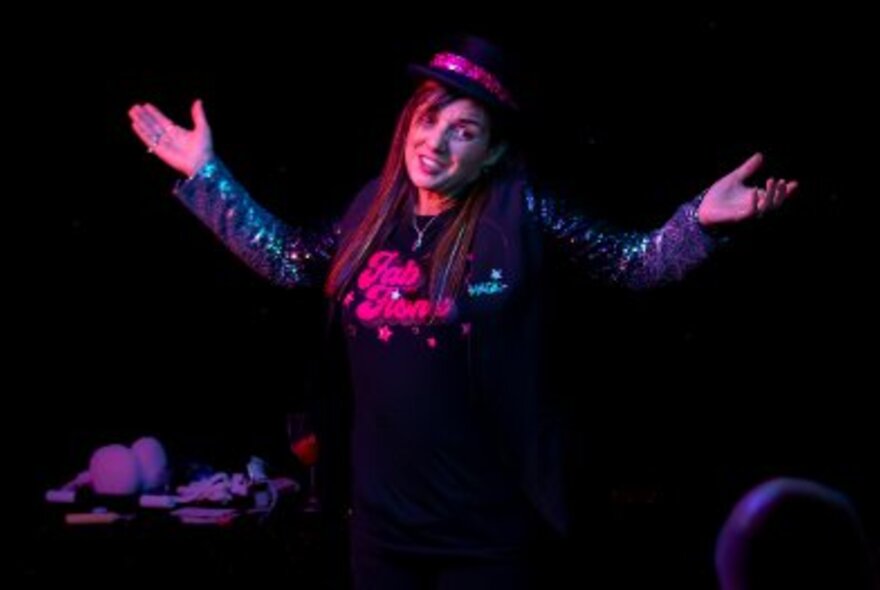 Woman wearing a black hat with pink ribbon performing on a stage, her arms open wide.