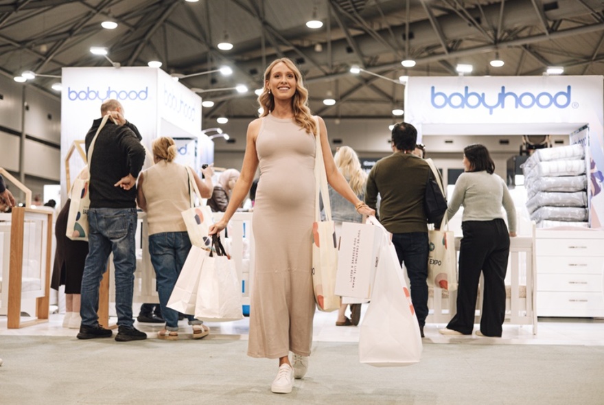 A pregnant woman holding multiple carrier bags at an expo with people standing around product stands.