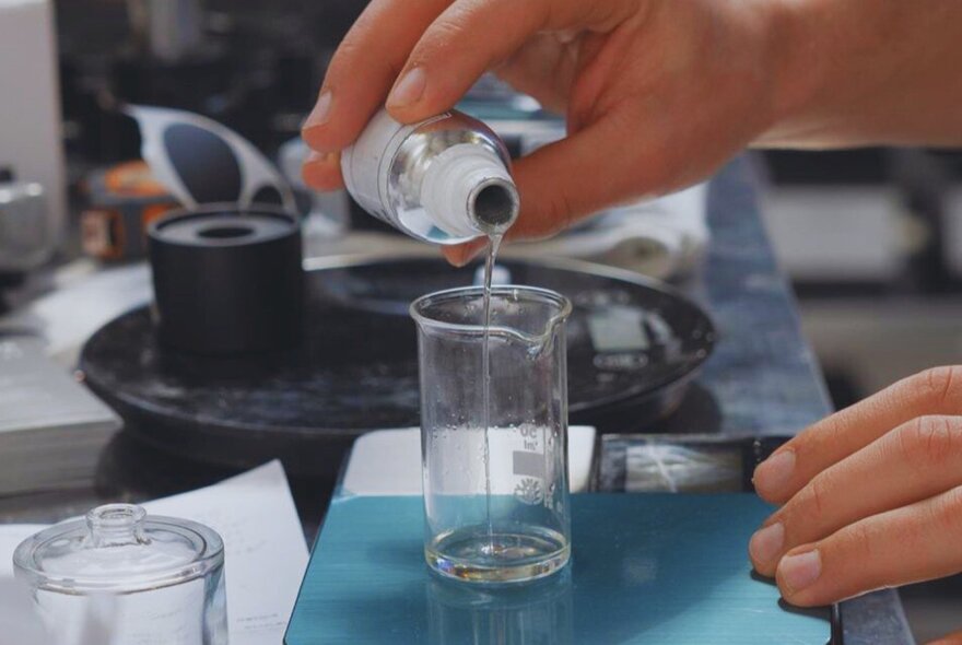 A hand pouring liquid from a small bottle into a small beaker, at a perfume mixing workshop.