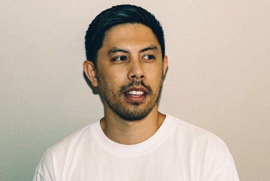 Comedian Chris Nguyen, a young man with short dark hair, his head turned slightly to one side with his eyes averted, wearing a white t-shirt. 