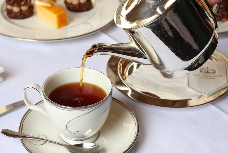 A fine china tea cup and saucer, being filled by a stainless steel teapot on a white clothed table with high tea delicacies in the background.