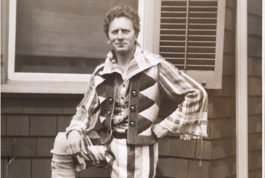 Black and white photo of Percy Grainger wearing a shirt with open flared sleeves, a patterned vest and striped trousers, standing in front of a house.