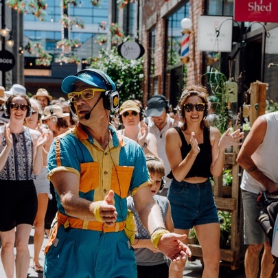 A musical walking tour being led through a Melbourne laneway by a man wearing a blue cap, headphones, speaker and overalls.