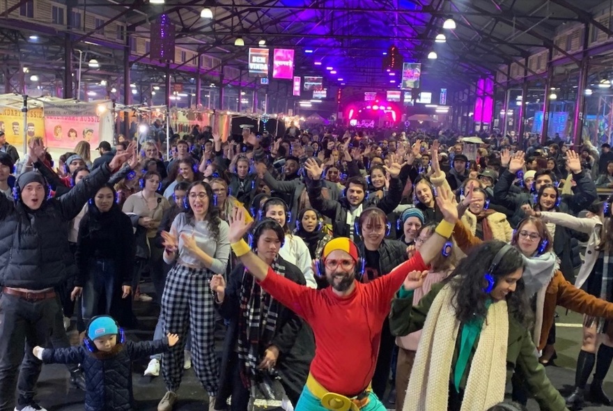 Guru Dudu leading a crowd of walkers and dancers in an indoor shed at Queen Vic Market at night.