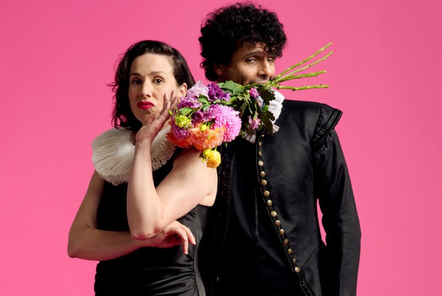 Actors wearing black and white Shakespearean-era clothing with ruffs, the woman holding her chin in her hand, the male actor with a boquet of flowers in his mouth. 