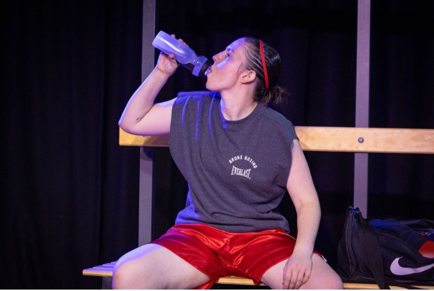 A scene from the play Bitch Boxer, featuring a performer in red boxing shorts and a dark sleeveless top resting on a locker room bench.