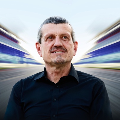 Guenther Steiner, a middle-aged man with short hair wearing a black shirt, centred against a stylized, highly-blurred background featuring rapid motion lines that mimic a Formula 1 car speeding down a racetrack.