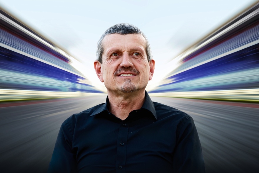 Guenther Steiner, a middle-aged man with short hair wearing a black shirt, centred against a stylized, highly-blurred background featuring rapid motion lines that mimic a Formula 1 car speeding down a racetrack.