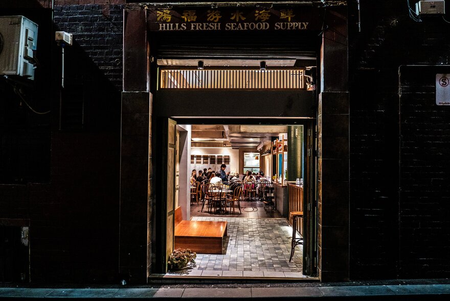 A small entry to a Chinese restaurant in a laneway. There are people dining inside and a sign above the door frame that says Hills Fresh Seafood Supply. 