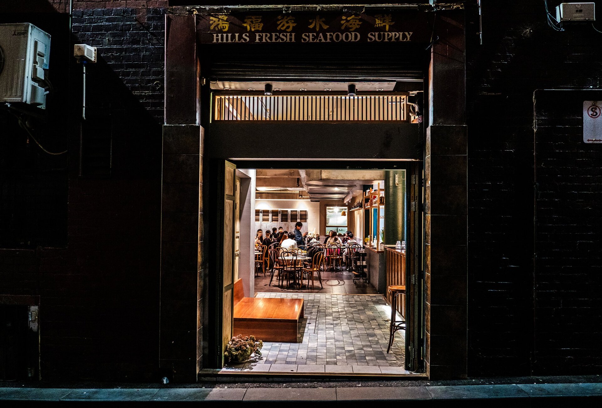 A small entry to a Chinese restaurant in a laneway. There are people dining inside and a sign above the door frame that says Hills Fresh Seafood Supply.