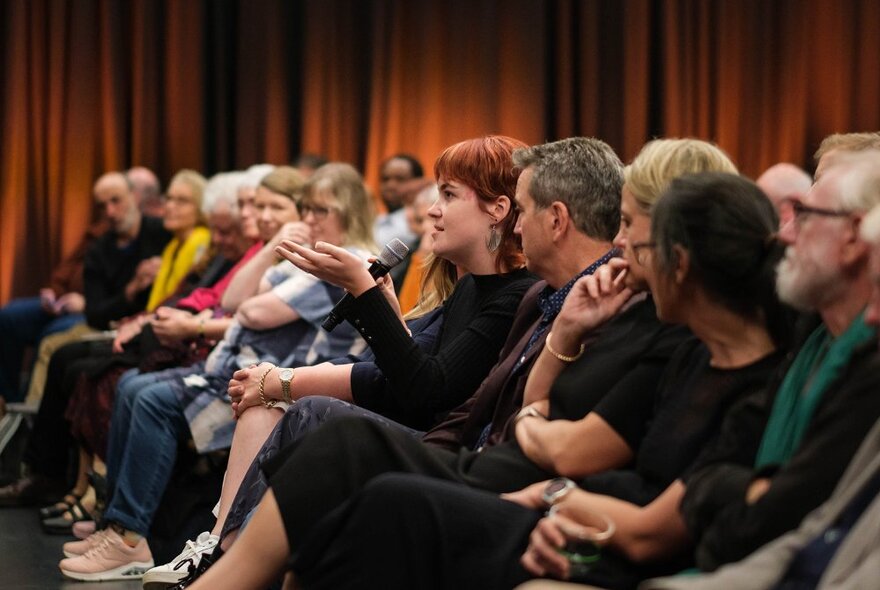 An audience in a theatre setting with the lights on and one woman talking and gesticulating towards the front. 