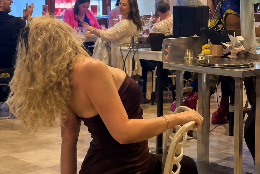 A woman in a strapless top seated backwards on a chair arching her back so her curly blond hair is hanging down behind her.