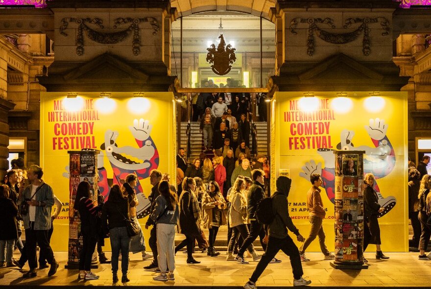 The Melbourne Town Hall entrance with bright comedy festival signage and people walking past, and lined up on a staircase.