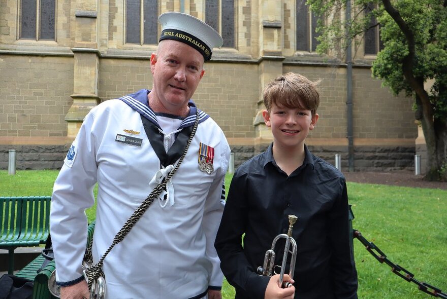 A man in an Australian Navy uniform stands next to a young boy with a bugle outside a church.