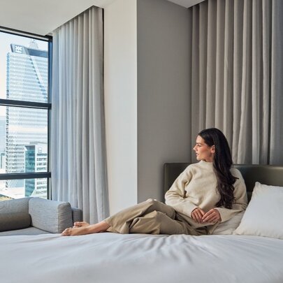 A woman relaxing against a bedhead in a hotel room, and gazing out the floor to ceiling window at the view of the city. 