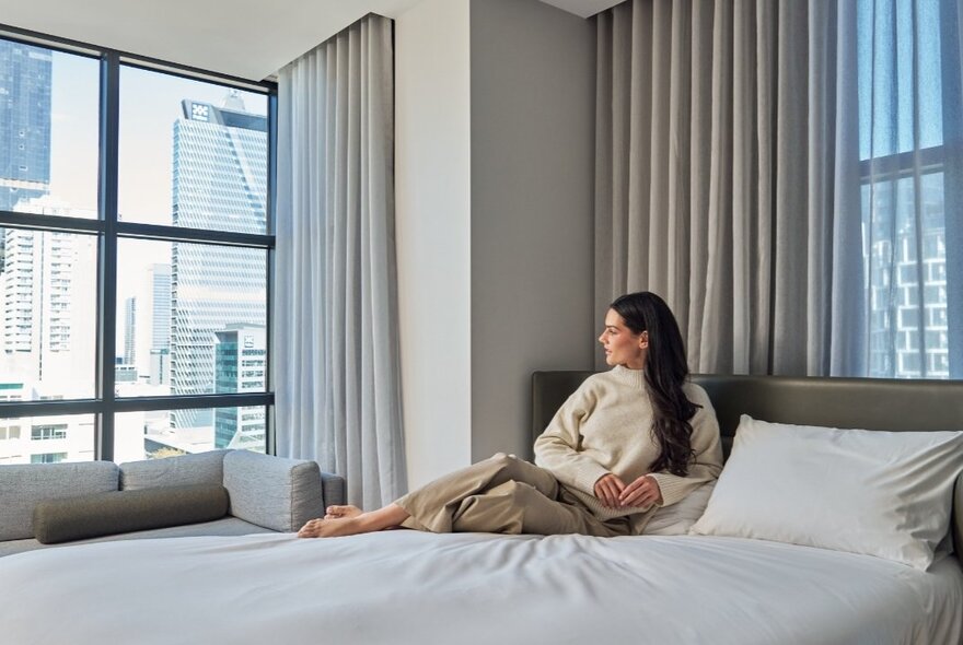 A woman relaxing against a bedhead in a hotel room, and gazing out the floor to ceiling window at the view of the city. 