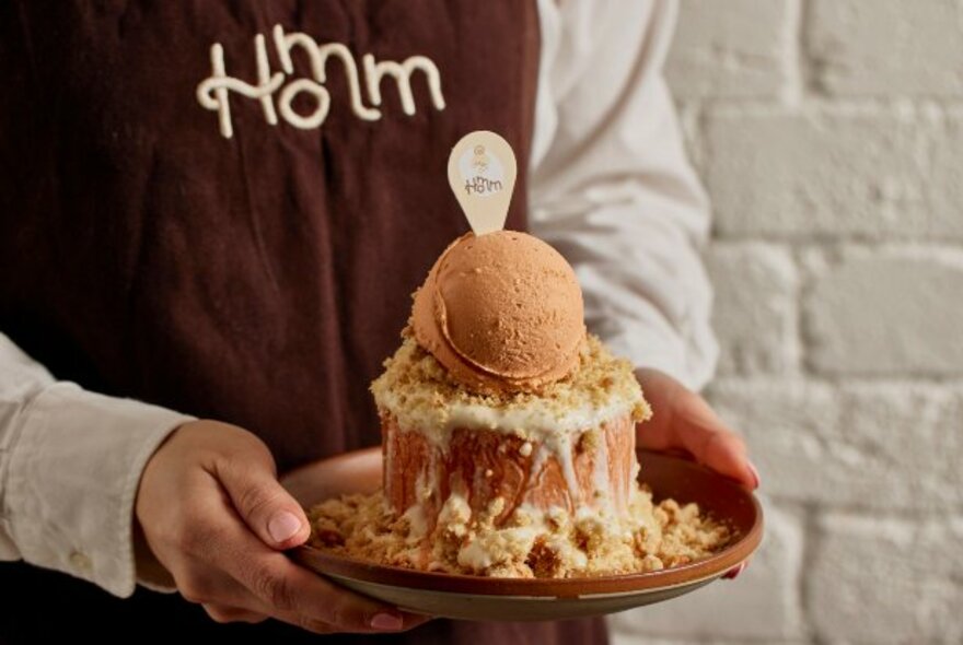 A person in a brown apron holding a plate with an elaborate dessert on it with a white brick wall in the background. 