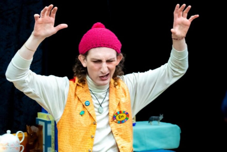 Performer wearing a yellow waistcoat and red hat, seated with arms raised.