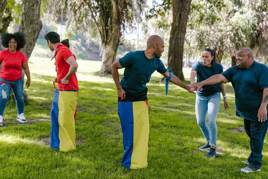 A group of people doing a team activity in a leafy park setting, with two individuals' legs wrapped in a blue and yellow fabric.