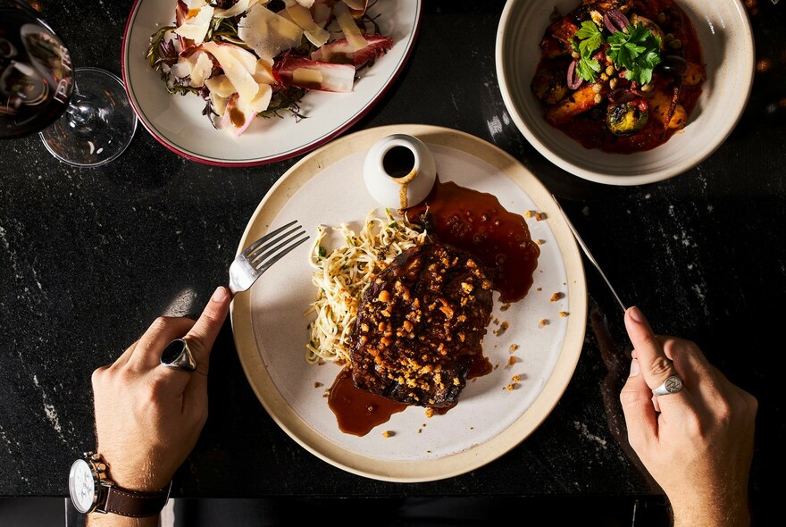 Hands holding cutlery about to cut into a plateful of steak, with accompanying dishes and wine glass.