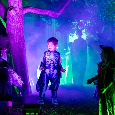 Three children in neon-lit Halloween costumes in a creepy Halloween scene lit up with purple and green neon lights.
