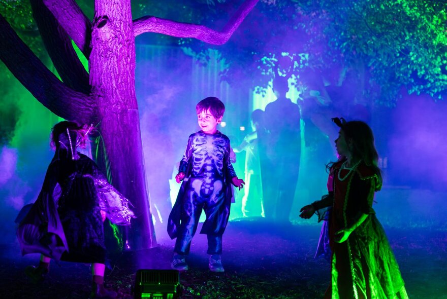 Three children in neon-lit Halloween costumes in a creepy Halloween scene lit up with purple and green neon lights.