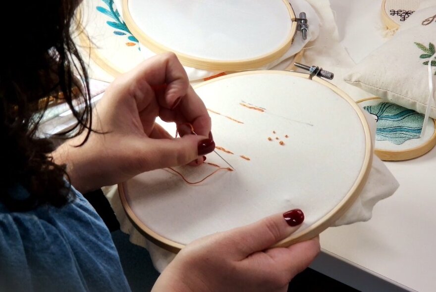A hand holding a needle and thread and embroidering a stretched piece of cotton cloth.
