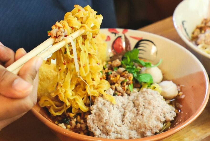 Hands holding chopsticks and Thai noodles from a bowl with rice.