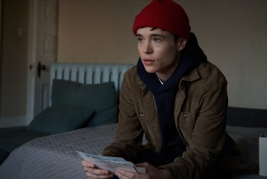 A still from a film of a person wearing a red beanie and looking pensive, sitting on the edge of a bed and holding papers in their hands.