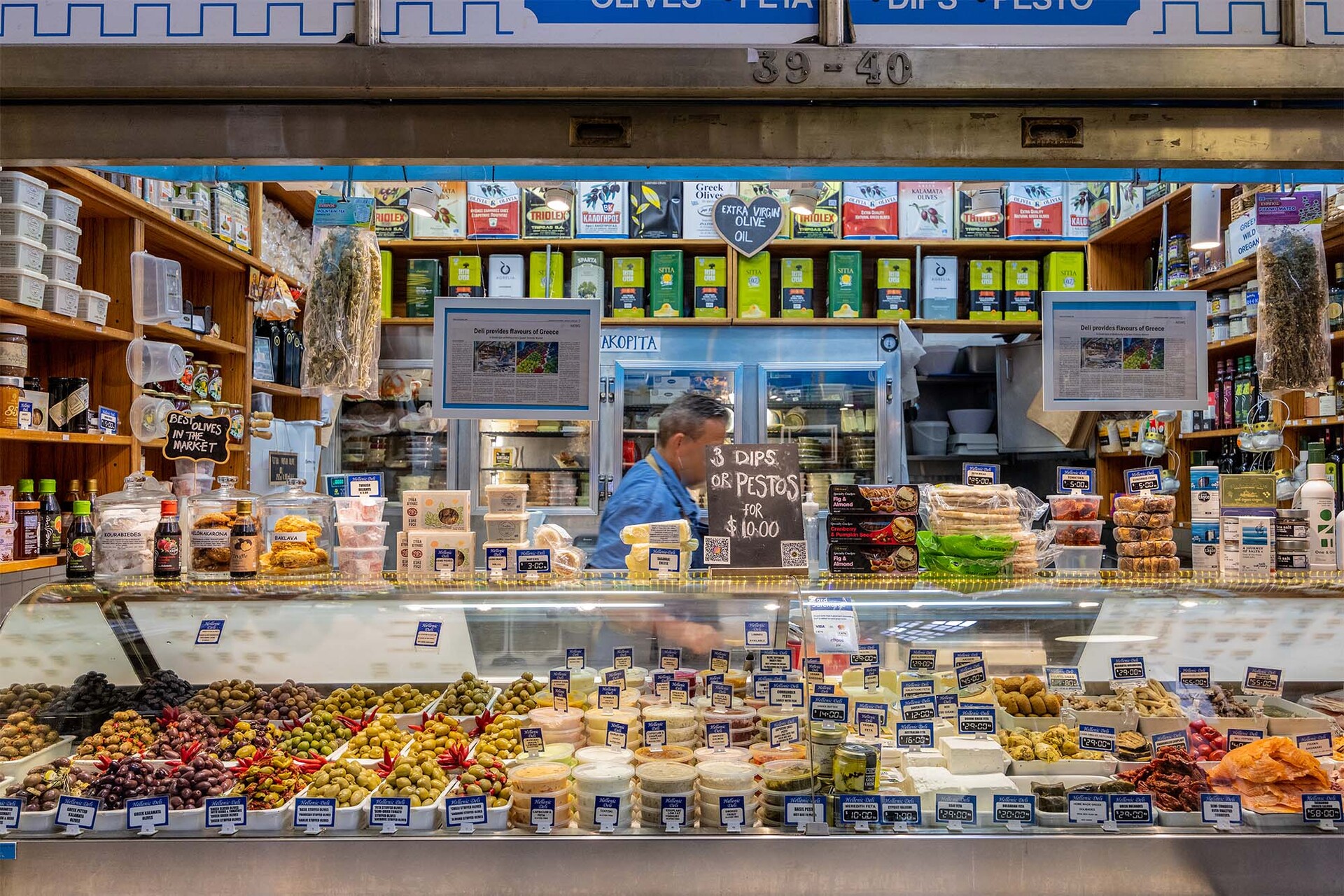 A man is behind the counter at a Greek deli which is packed with dips, olives and other goods.