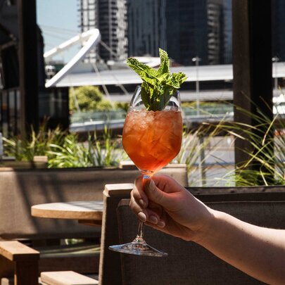 A hand holds up an orange-red cocktail garnished with fresh mint on an outdoor terrace, with a waterfront and buildings visible in the background