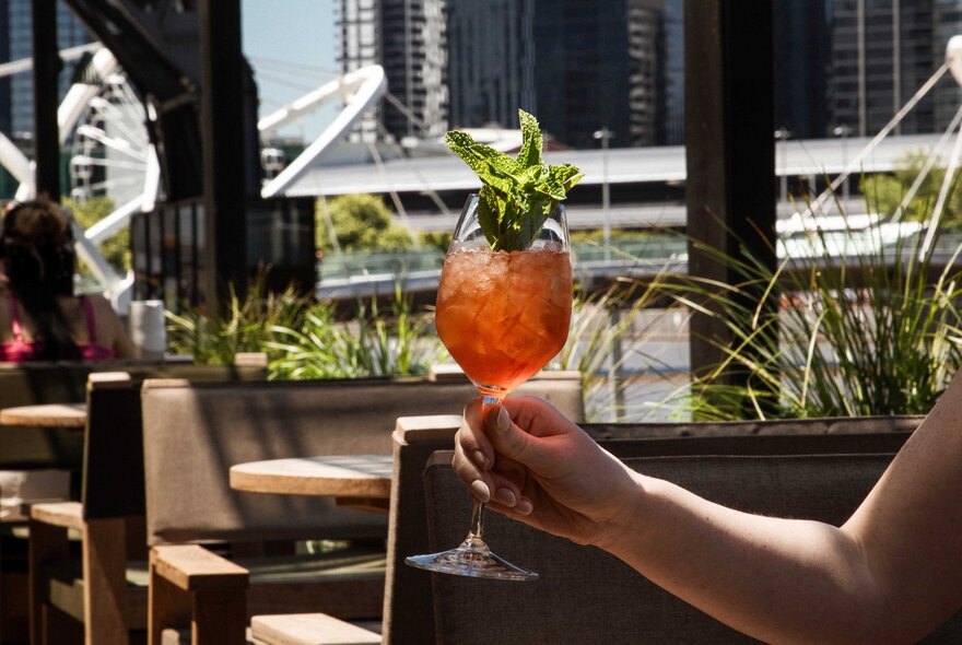 A hand holds up an orange-red cocktail garnished with fresh mint on an outdoor terrace, with a waterfront and buildings visible in the background