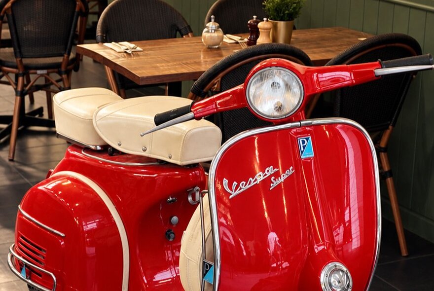 A close-up of a glossy red vintage Vespa scooter parked inside a restaurant, with empty wooden dining tables and chairs blurred in the background.