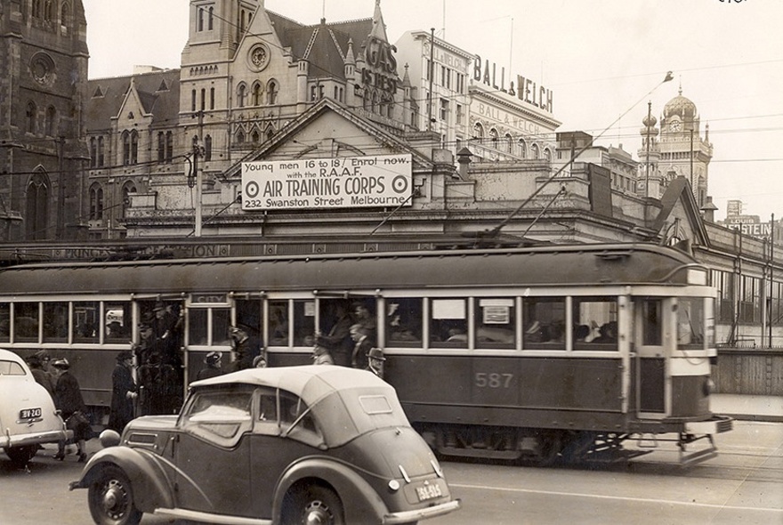 Black and white historical photograph of a vintage W class tram and a classic 1940s car on a busy street in front of the old Princes Bridge Station in Melbourne.