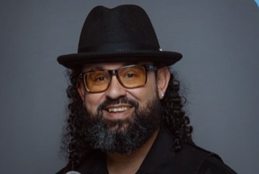 Portrait shot of Torres Strait Islander comedian, Leon Filewood, wearing a black fedora and glasses and smiling at the camera; grey background.