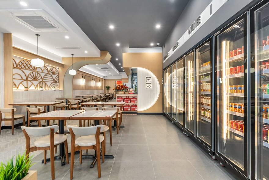 Cafe interior with dining tables and chairs next to a wall of drinks fridges under grey ceiling with lights.