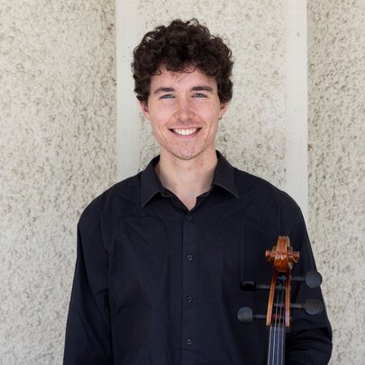 Cellist Boudewijn Keenan smiling against a stucco wall while holding his instrument. 