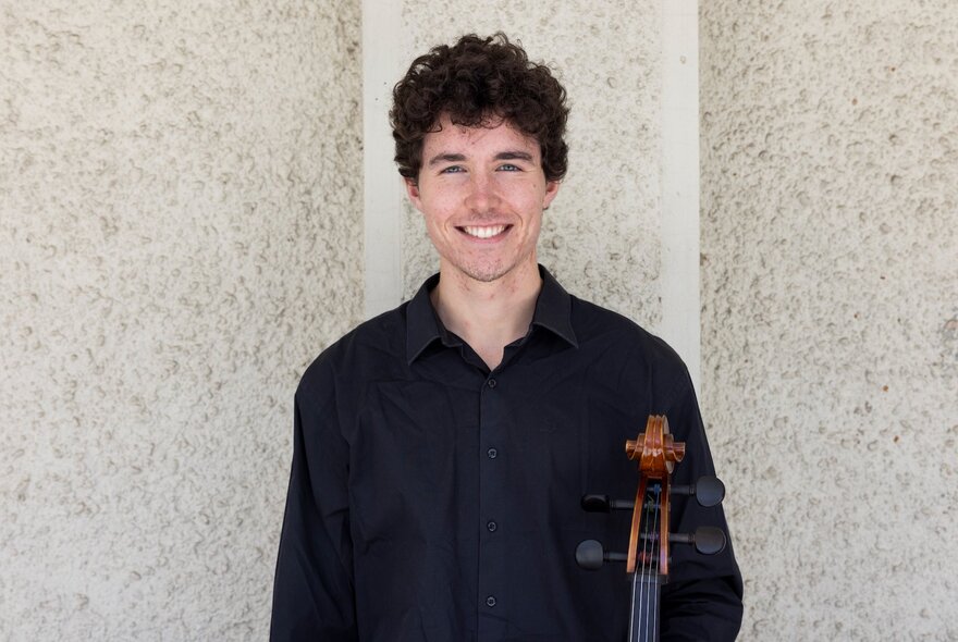 Cellist Boudewijn Keenan smiling against a stucco wall while holding his instrument. 