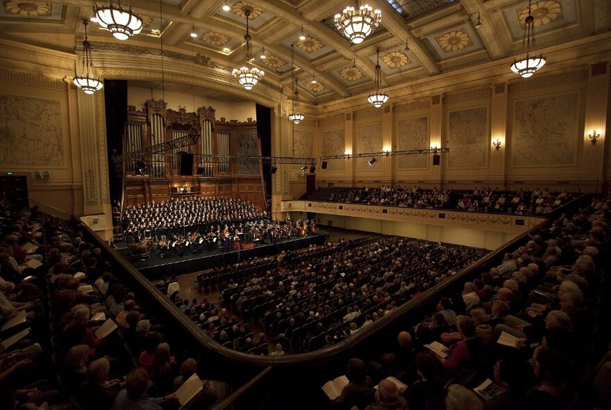 Interior of a grand hall, with a group of orchestra musicians and choir singers on a stage, a grand organ behind them, and people in seats listening to the concert being performed.