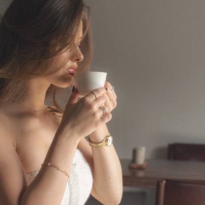 Model wearing bracelet, watch and rings, holding a cup to her face, wearing a strapless white tight-fitting top.