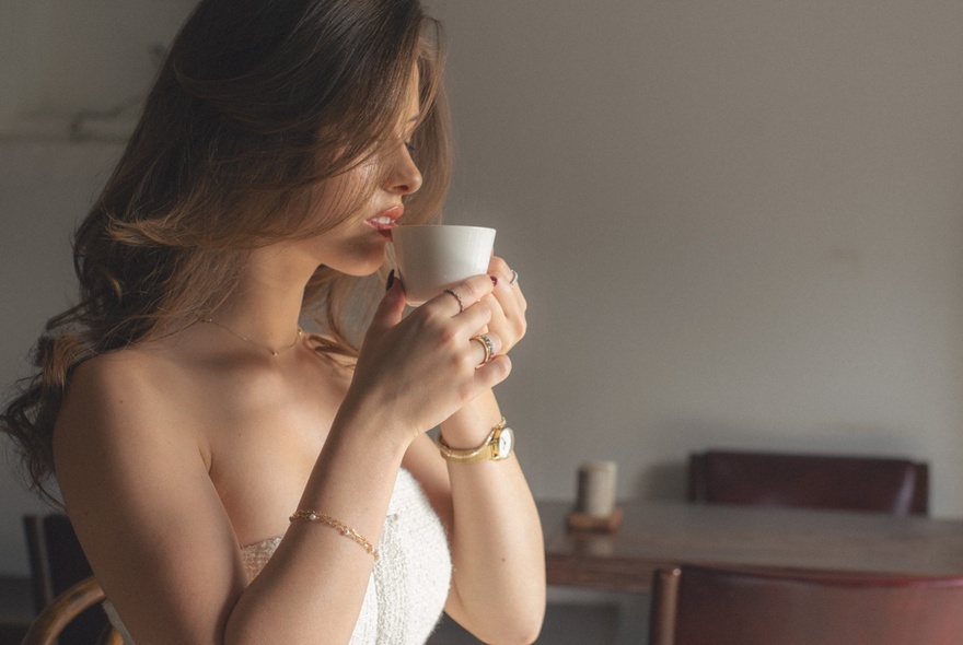Model wearing bracelet, watch and rings, holding a cup to her face, wearing a strapless white tight-fitting top.