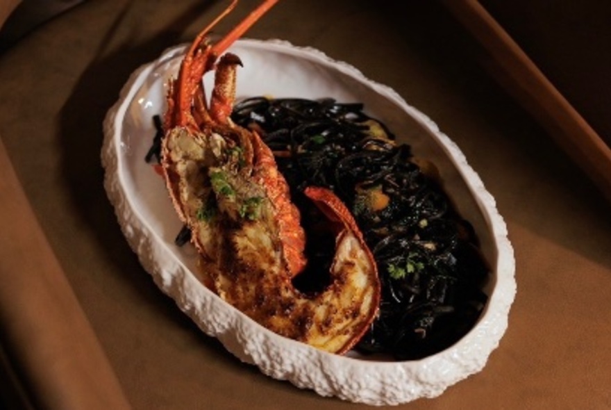 Large oval dish of half a filled lobster alongside black noodles, on a dining room table.