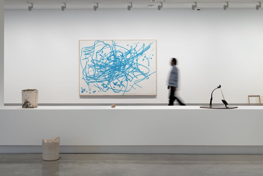 A large white room in a gallery, with a person in the background walking past a large contemporary artwork hanging on the wall.