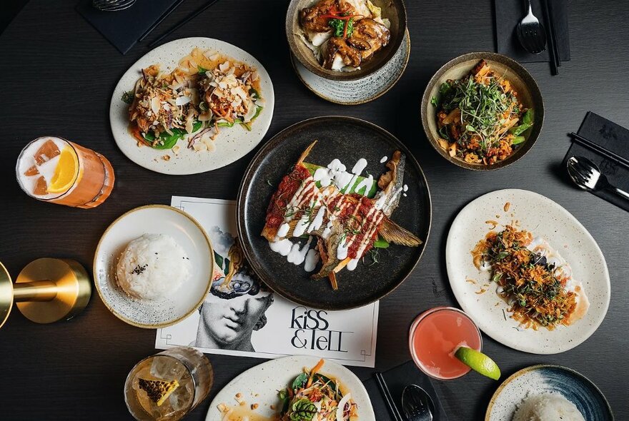 Overhead view of a black dining table in a restaurant filled with several dishes of food and cocktails.