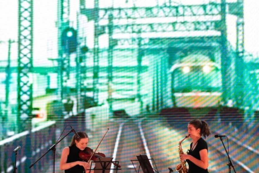 Violinist and saxophonist performing in front of a large screen displaying a trainline and train in Japan.