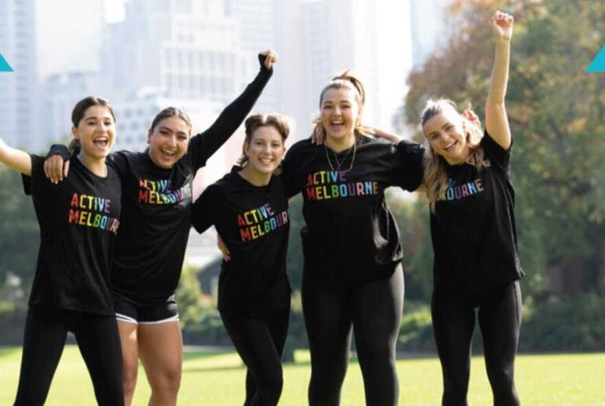 Five smiling girls wearing active wear, and matching t-shirts with the slogan ACTIVE MELBOURNE printed on them, in a group huddle outdoors in a park with their arms raised in the air.